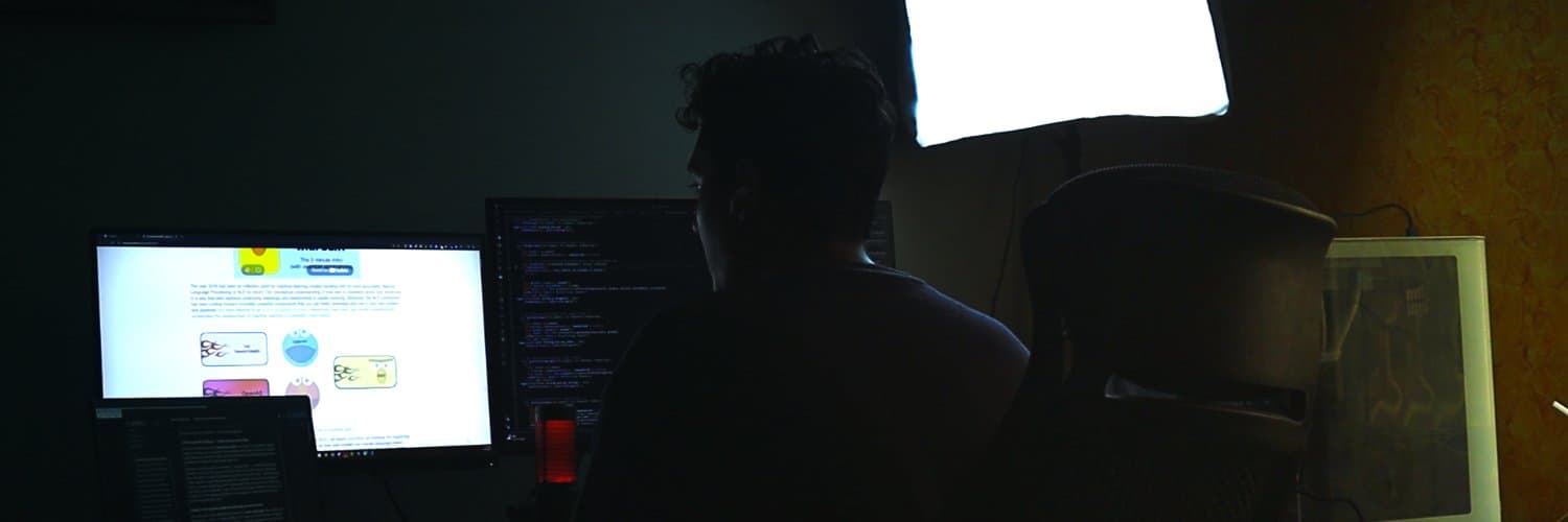 Muzammil at his desk, working across three monitors in a dark room.